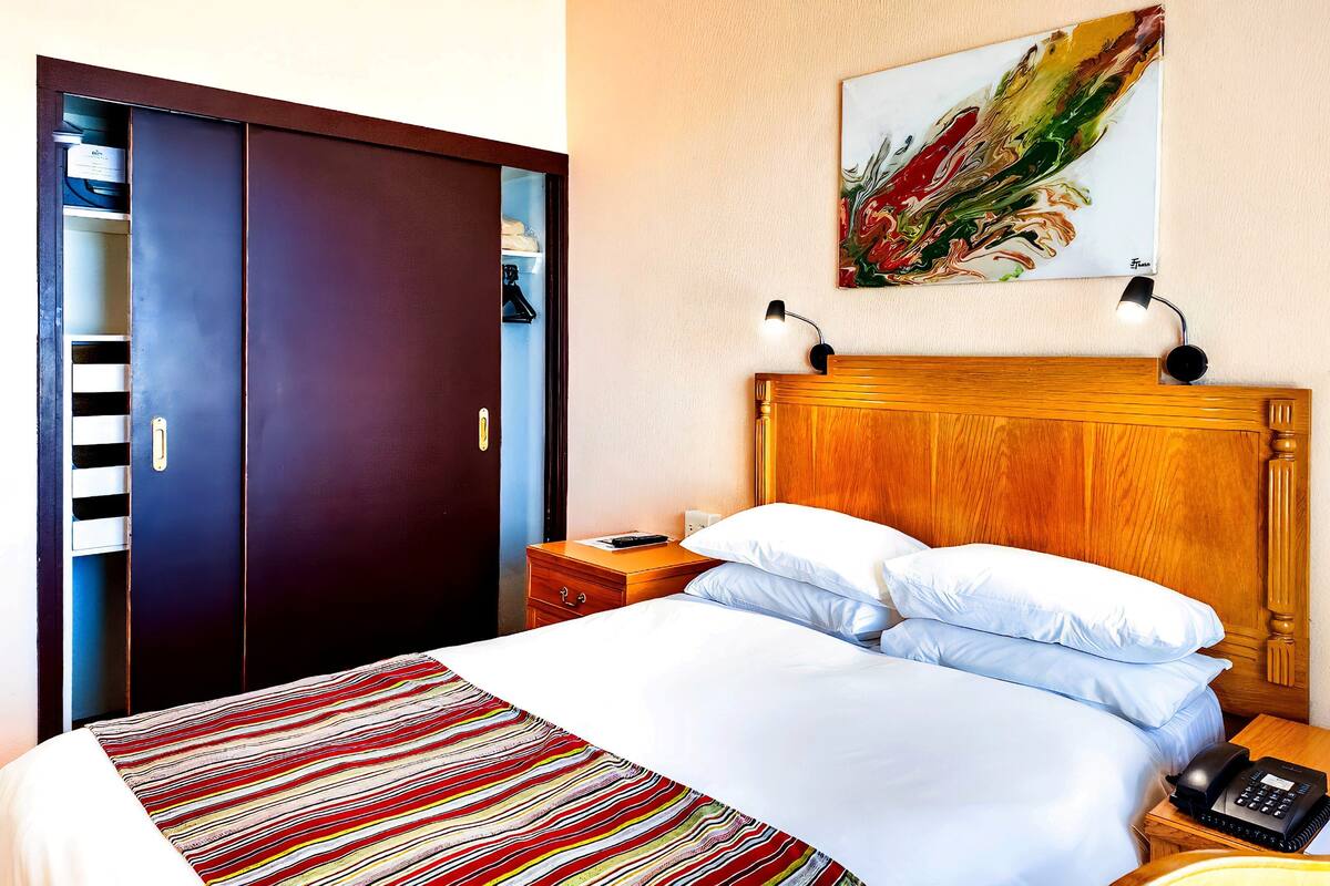 Une chambre d’hôtel avec un lit, une table de chevet, un téléphone, un tableau au mur et une armoire.
