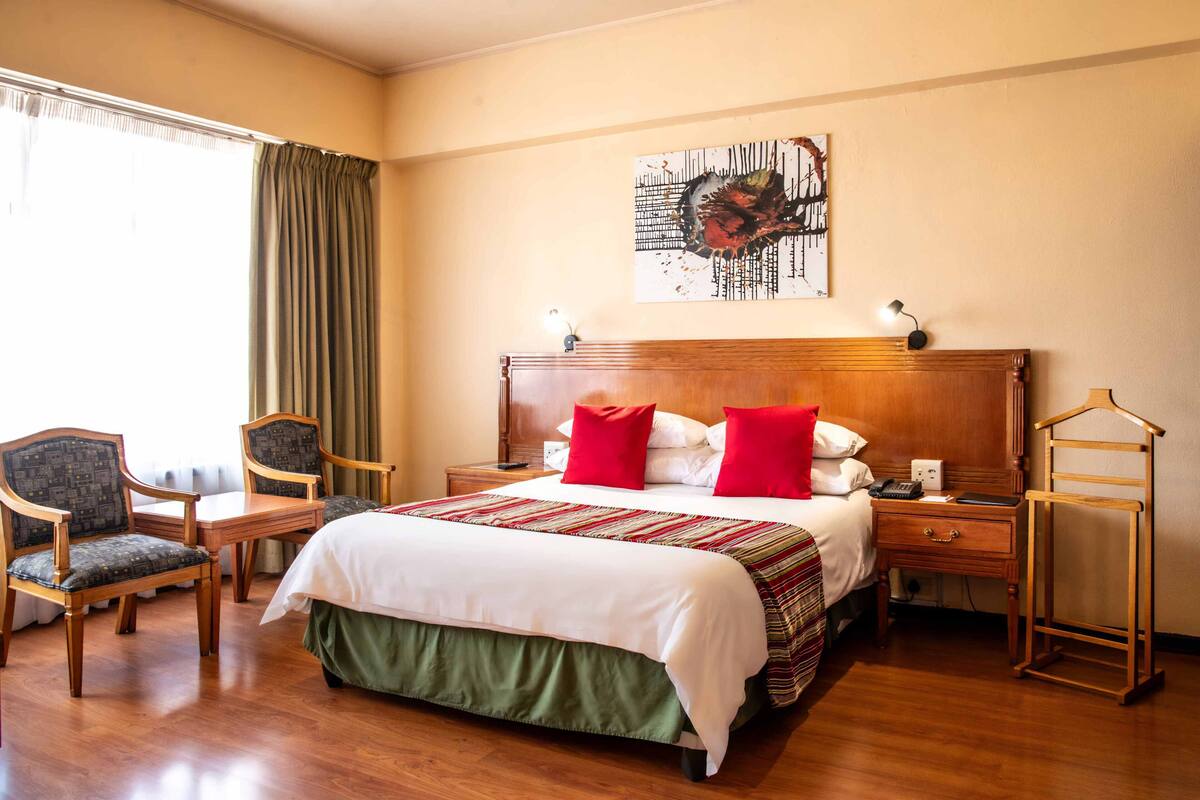 Une chambre d’hôtel avec un grand lit, deux chaises, une table de chevet, une commode en bois et un tableau au mur.