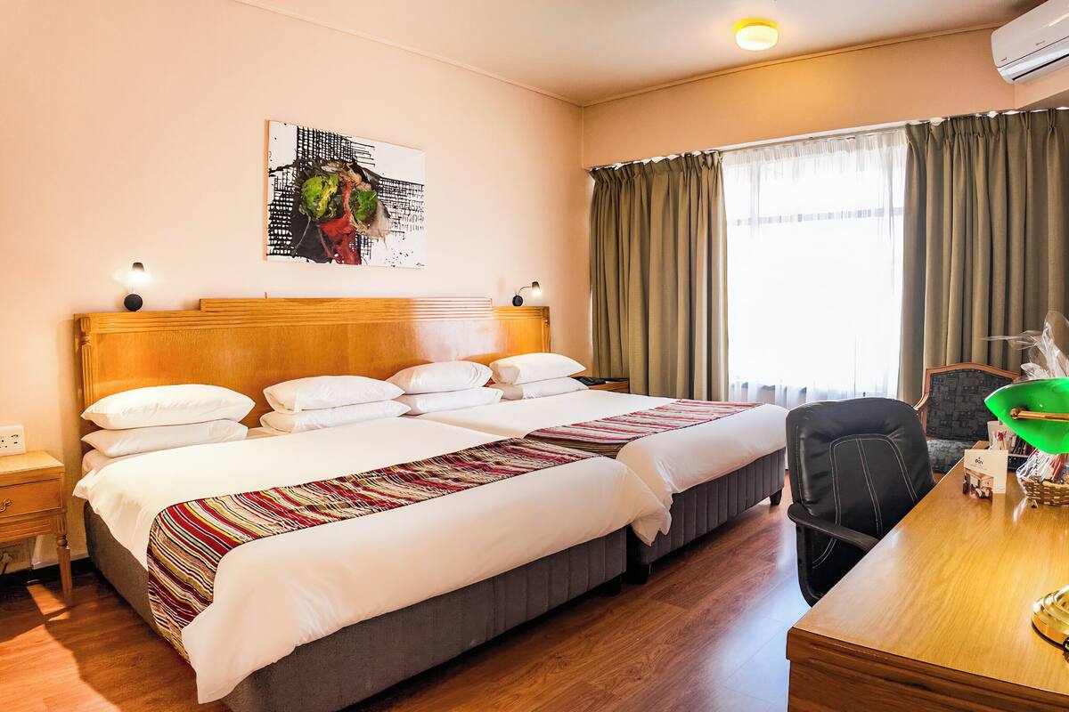 Une chambre d’hôtel avec un grand lit, un bureau avec une lampe, une chaise, une fenêtre avec des rideaux et un climatiseur fixé au mur.