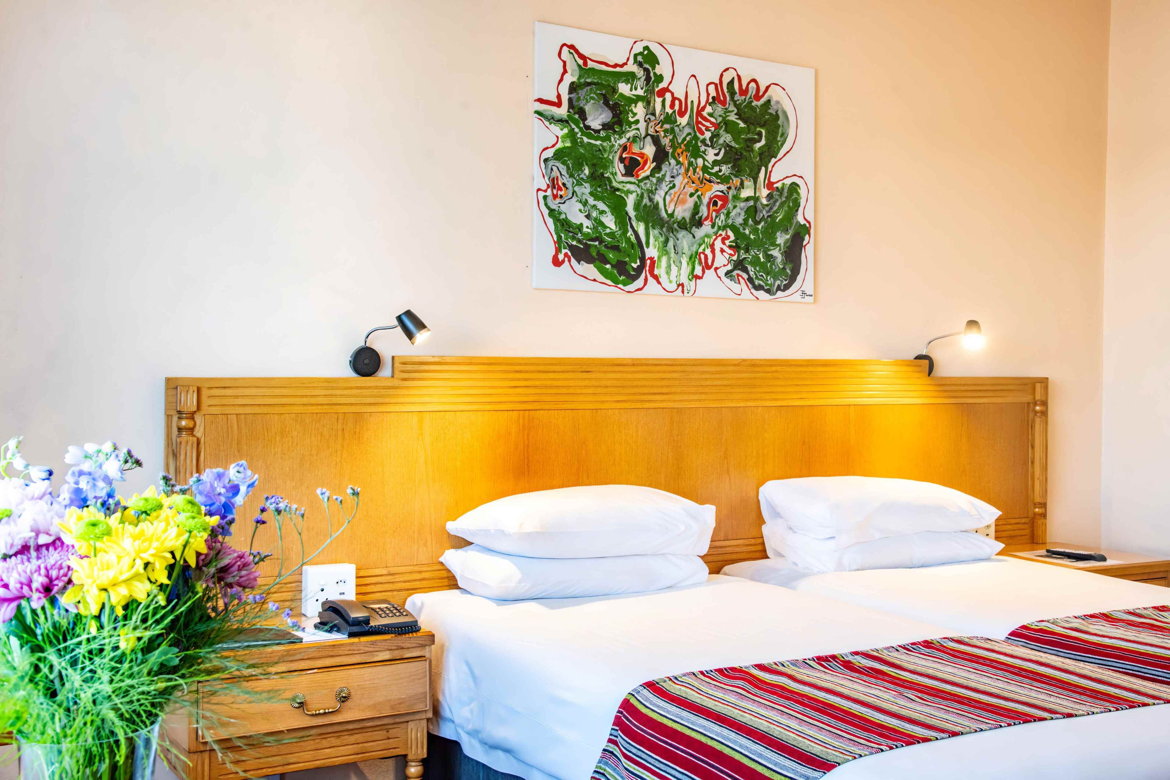 Habitación de hotel con cabecera de madera, cama con ropa de cama blanca, mesita de noche con lámpara, teléfono y arreglo floral.