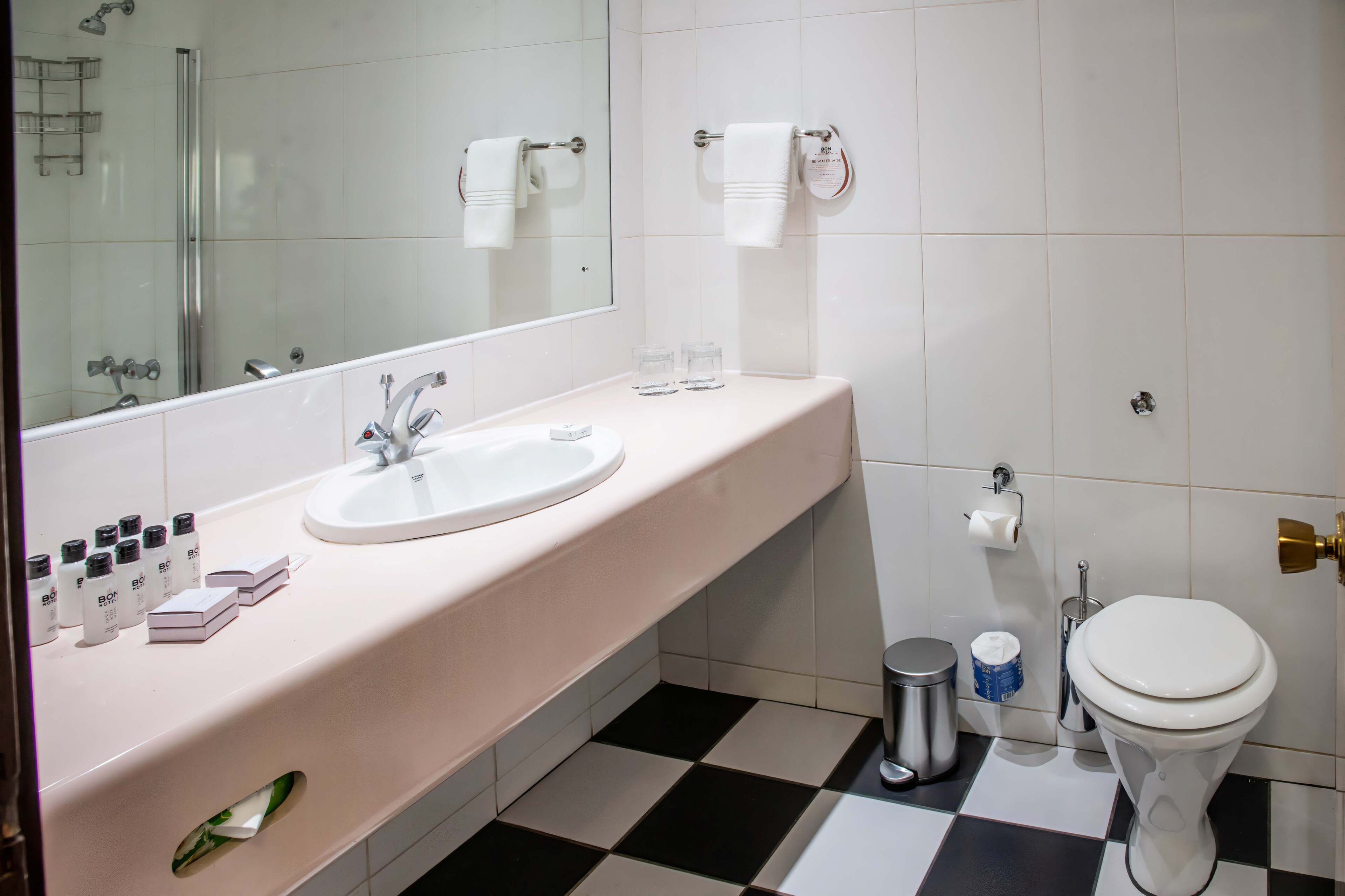 A bathroom with a white sink, mirror, and toilet. There are toiletries on the sink, a trash can, and a towel rack.