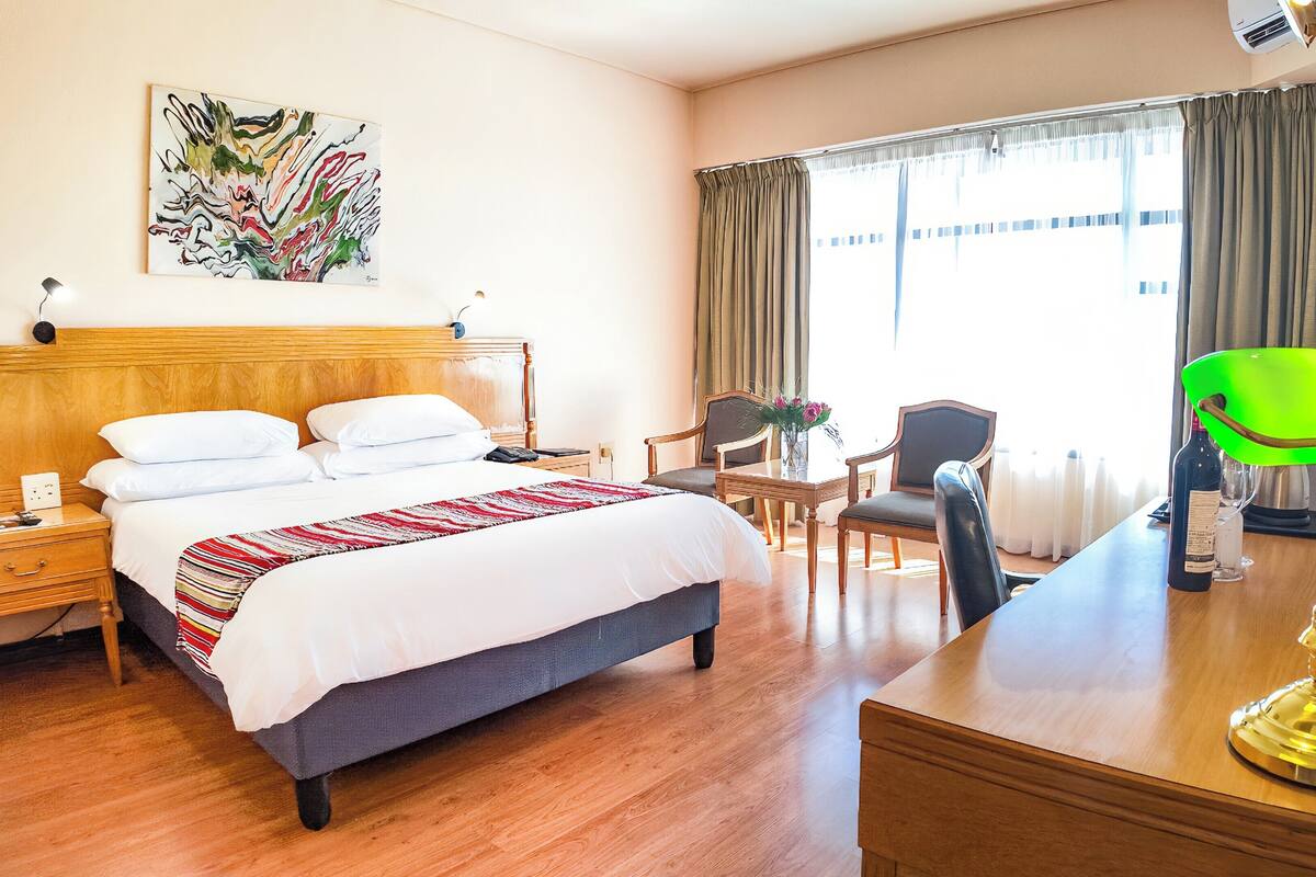 Une chambre d’hôtel avec un lit, un bureau, une chaise, une lampe et une fenêtre avec des rideaux.
