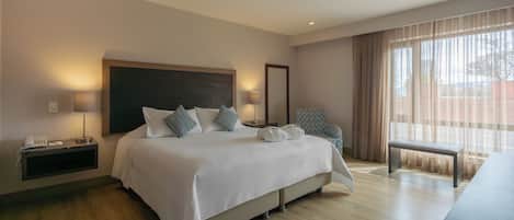 Una habitación de hotel con una cama grande, lámparas de noche, teléfono junto a la cama, una silla, un banco y una ventana con cortinas.