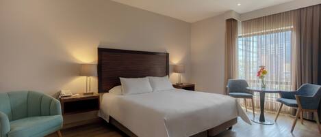 A hotel room with a bed, bedside tables, a lamp, a chair, a small table with a vase, and a window with curtains.