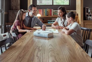 A family playing a board game at a wooden table with bookshelves in the background.