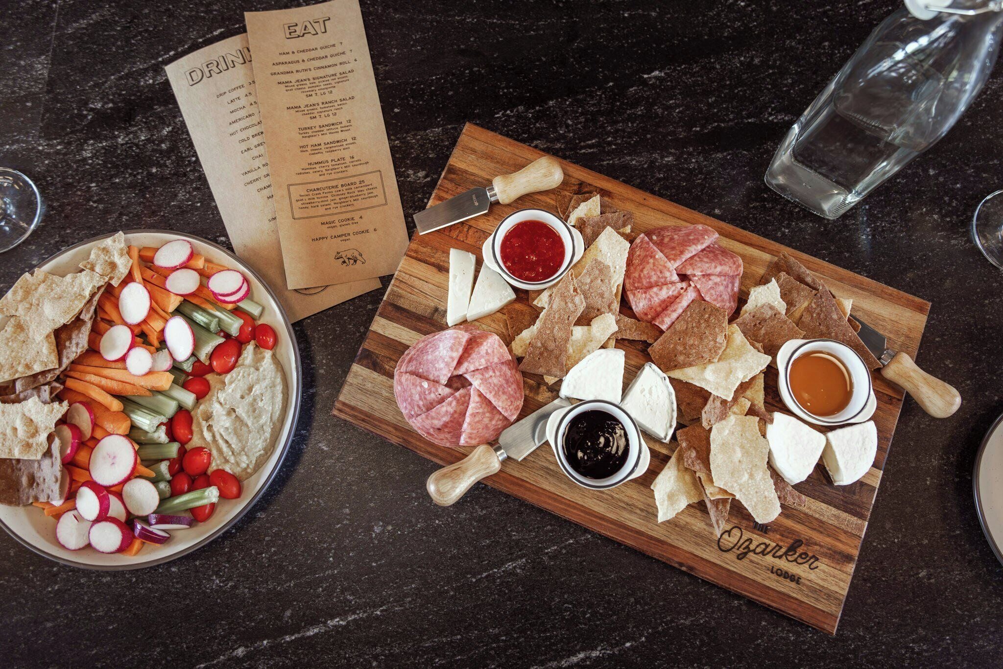A charcuterie board with various meats, cheeses, and vegetables on a wooden board, accompanied by a menu and a glass of water.