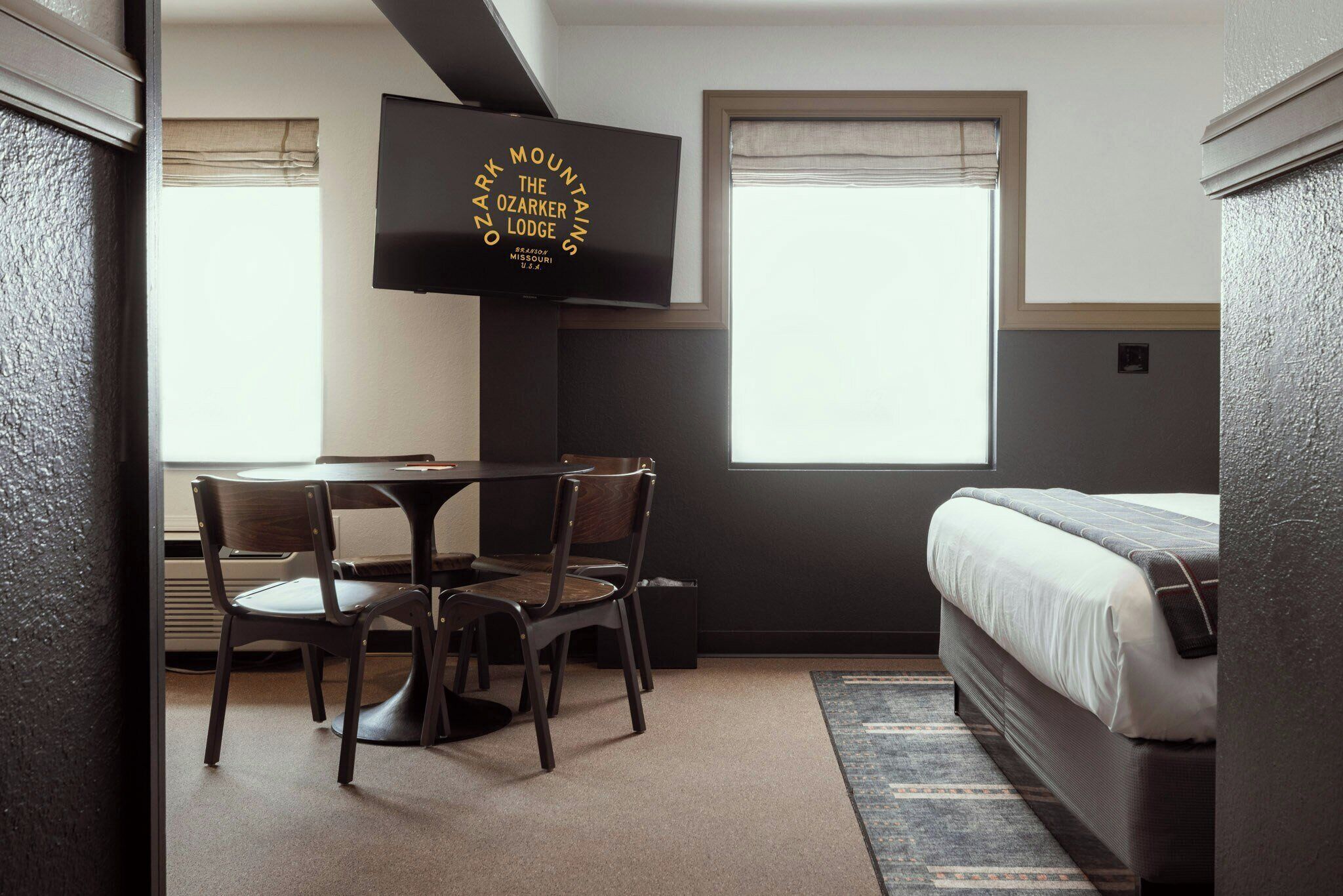 Una habitación de hotel con cama, televisor montado en la pared, mesa de comedor con sillas y ventana con persianas.