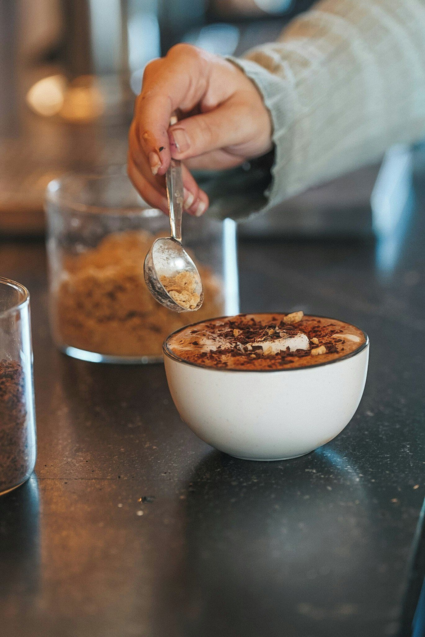 Une main tenant une cuillère au-dessus d’un bol de nourriture, avec un verre et un récipient en arrière-plan.