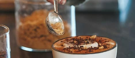 Une main tenant une cuillère au-dessus d’un bol de nourriture, avec un verre et un récipient en arrière-plan.