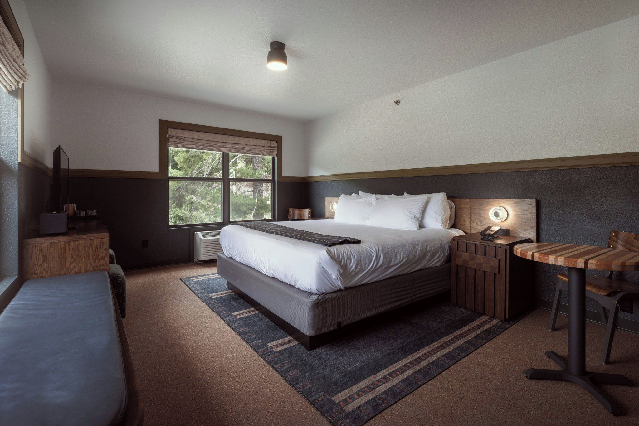 A hotel room with a large bed, a bedside table, a chair, and a window with a view of trees.