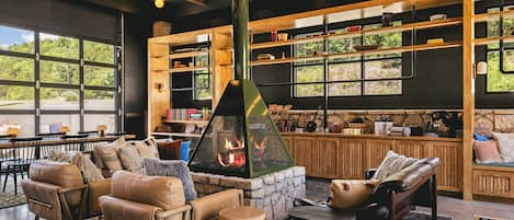 A cozy living area with leather sofas, a stone fireplace, wooden shelves, and large windows overlooking greenery.