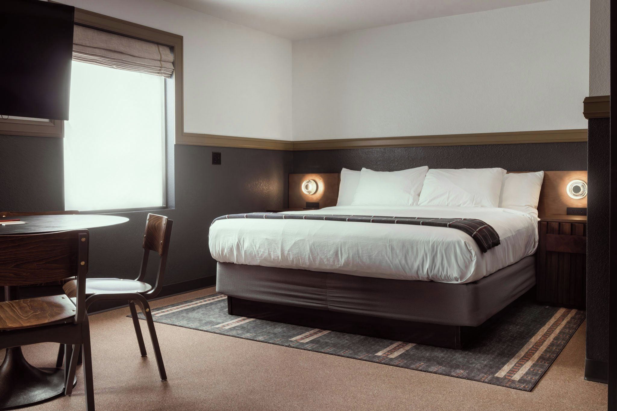 Une chambre d’hôtel avec un lit, des tables de chevet, une chaise, un bureau et une fenêtre avec des rideaux.