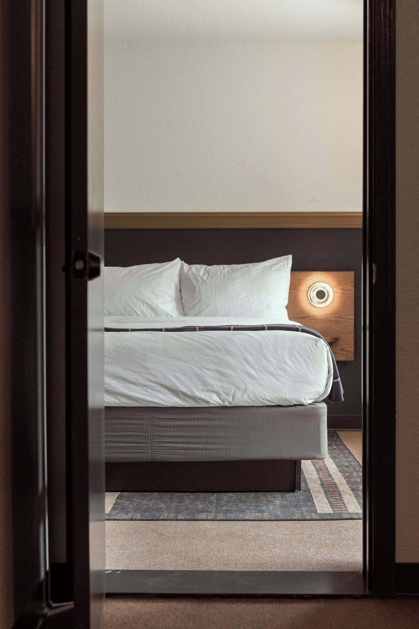 A hotel room with a bed, a headboard with a circular light, and a carpeted floor.