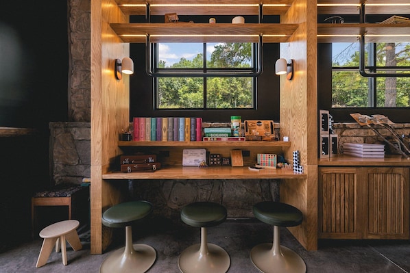 A cozy reading nook with a wooden bookshelf, a window with a view of trees, and three green bar stools.