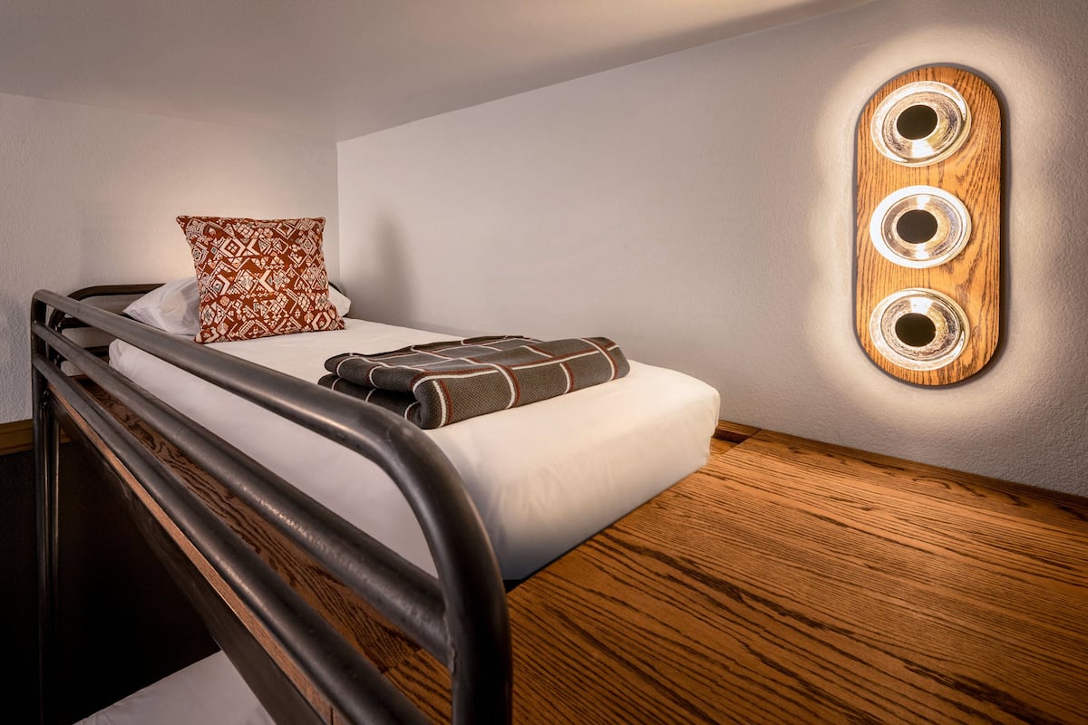 A single bed with a headboard, a patterned pillow, a blanket, and a wall-mounted light fixture.