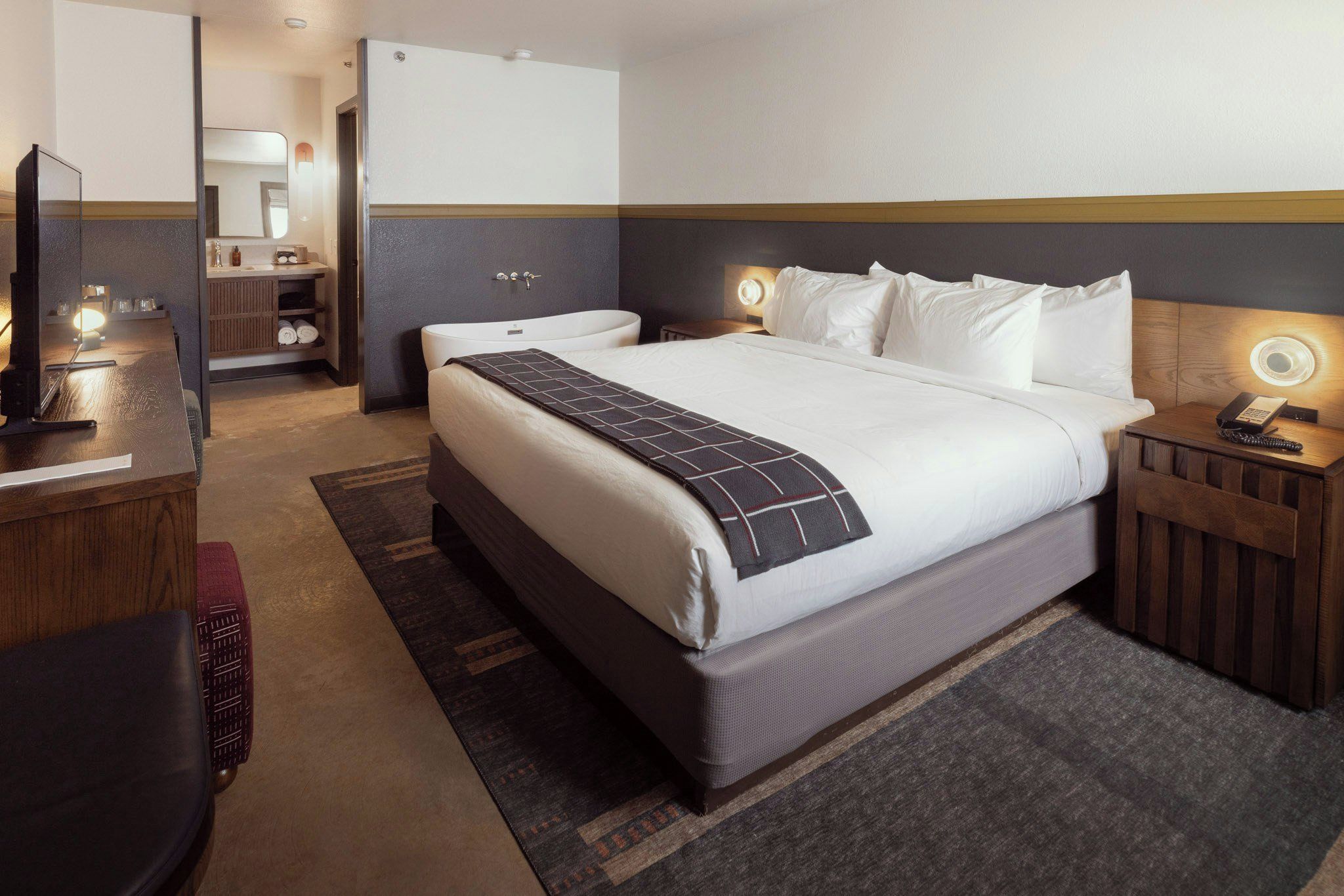 A hotel room with a large bed, bedside tables, a desk with a lamp, and a bathroom visible through an open door.
