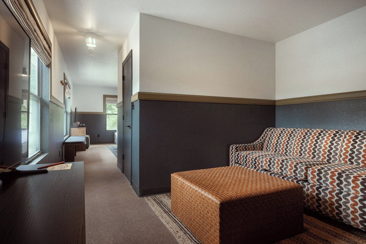 A modern hotel room with a dark wood floor, a brown upholstered sofa, a patterned rug, and a small wooden table with a lamp.