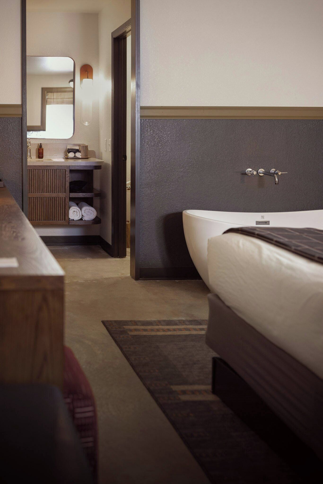 A hotel room with a bed, a bedside table, a mirror, and a bathroom visible through an open door.