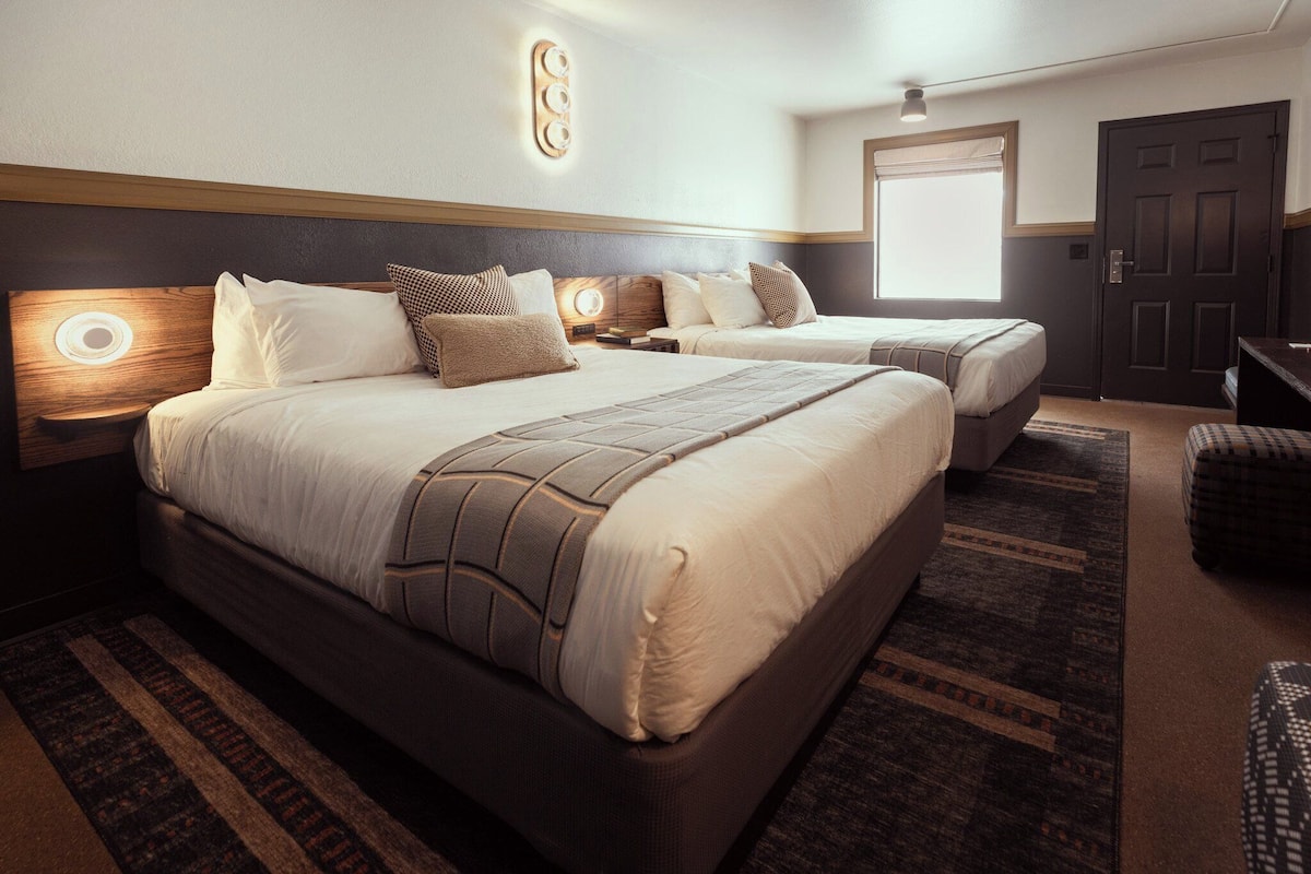 A hotel room with two beds, a large window, a door, and a wall-mounted light fixture.