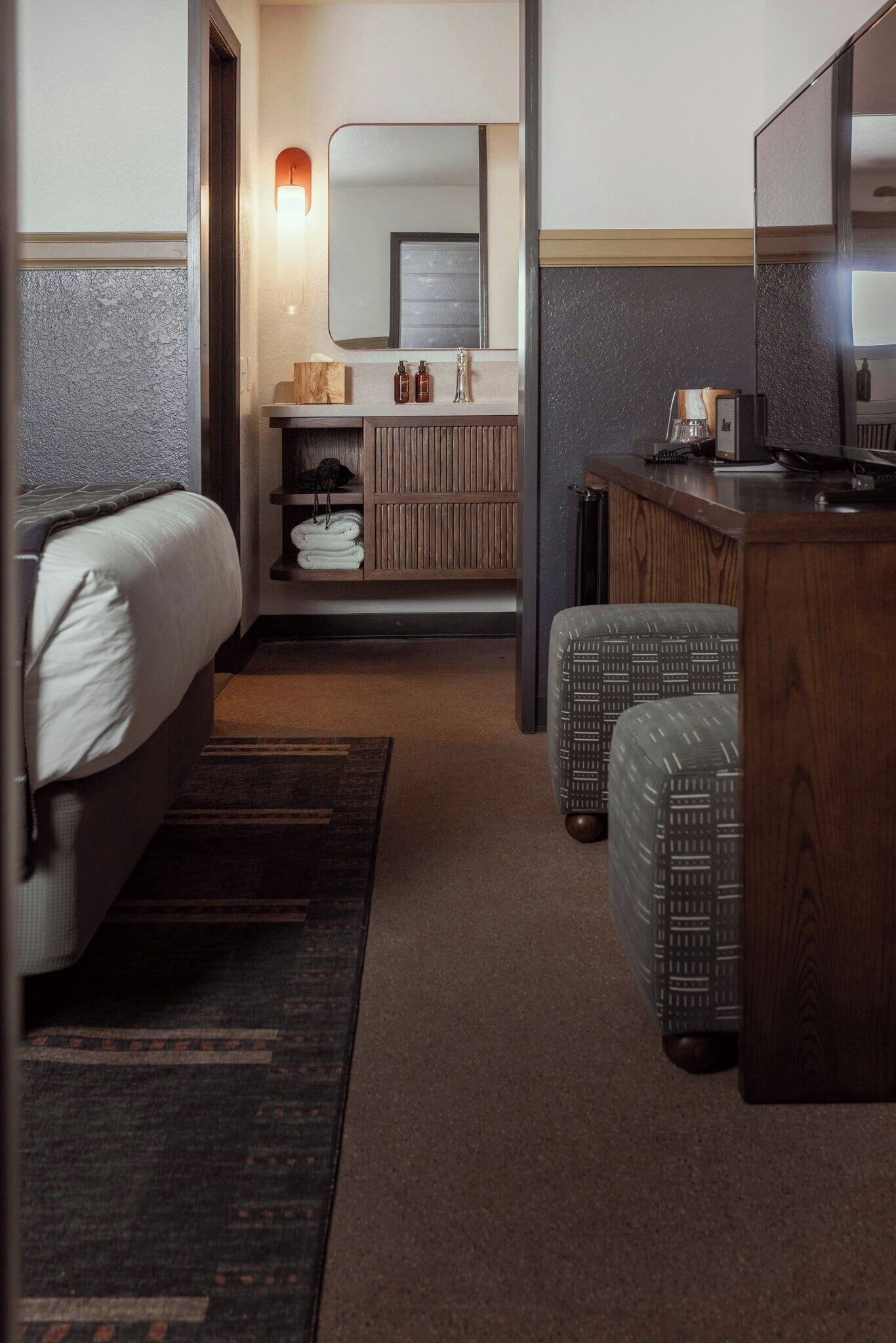 Habitación de hotel con cama, escritorio, espejo, pequeño rincón de descanso y baño con toallero.