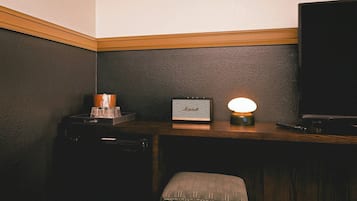 A corner of a room with a dark wall, a wooden desk, a small table with a lamp, a television, and a chair.