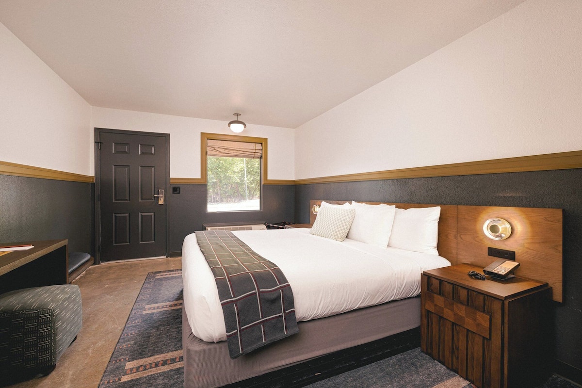 A bedroom with a large bed, a bedside table with a lamp, a desk, and a door.