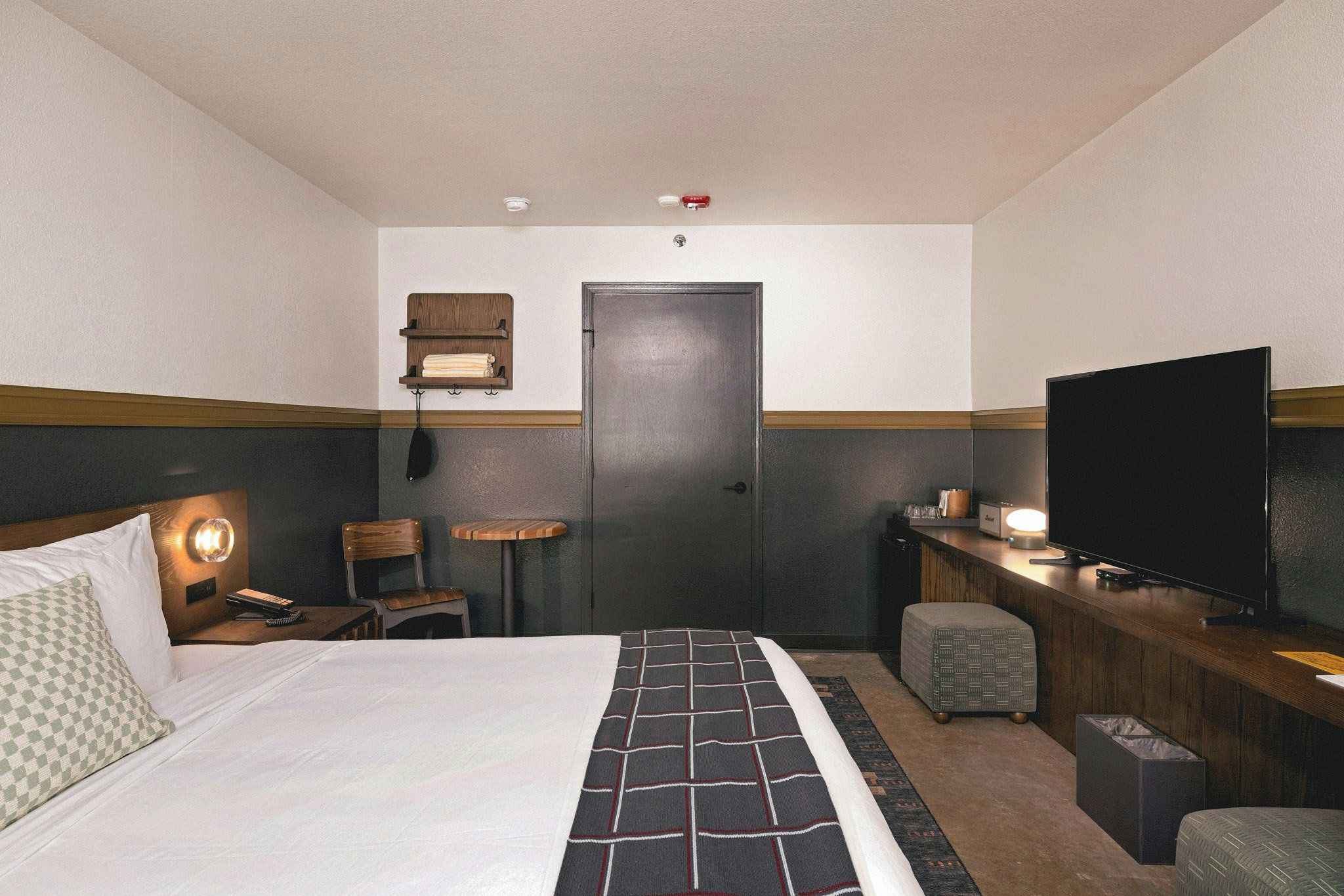 A hotel room with a bed, a desk with a television, a chair, a small table, and a wall-mounted shelf.