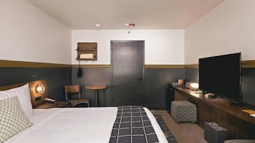 A hotel room with a bed, a desk with a television, a chair, a small table, and a wall-mounted shelf.