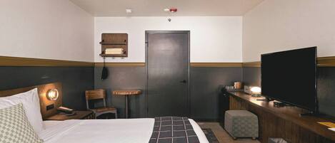 A hotel room with a bed, a desk with a television, a chair, a small table, and a wall-mounted shelf.