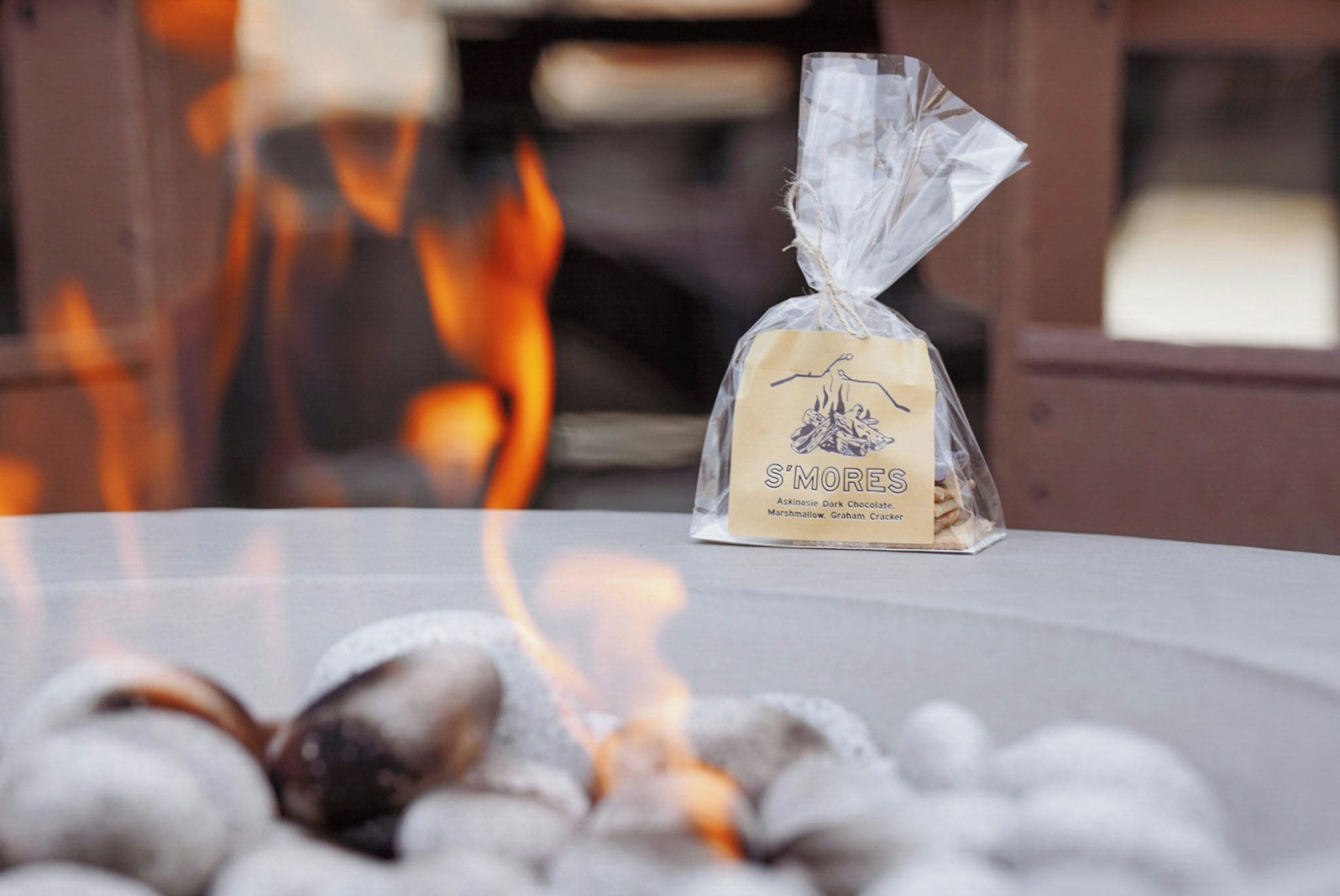 A bag of S'mores marshmallows, graham crackers, and dark chocolate on a table with a fire in the background.
