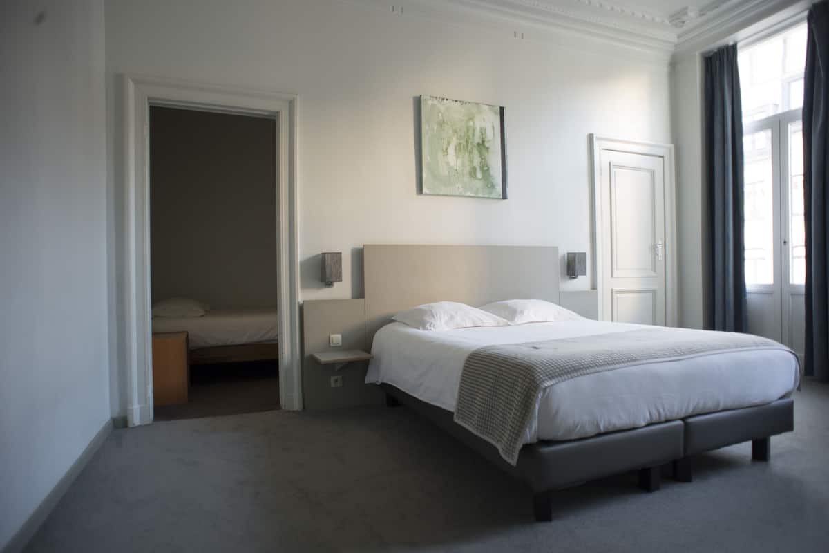 A bedroom with a large bed, a nightstand, a wall-mounted lamp, and a door leading to another room.