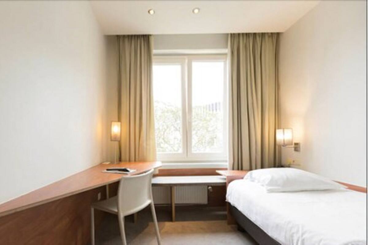 A hotel room with a bed, a desk, a chair, a window with curtains, and wall-mounted lamps.