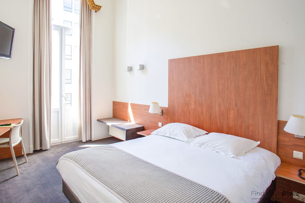 A hotel room with a large bed, a wooden headboard, a bedside table with a lamp, and a window with curtains.