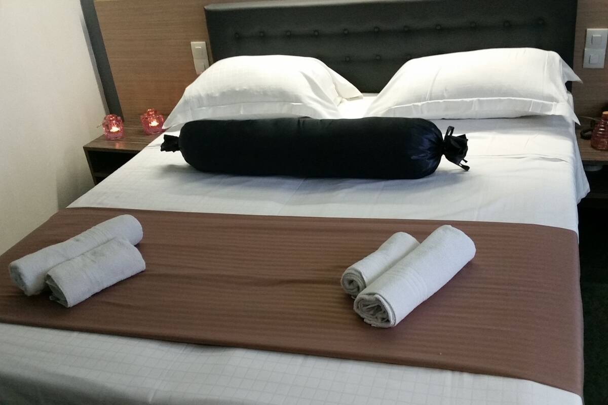 A neatly made bed with a brown bedspread, two rolled white towels, and a black pillow on the headboard.