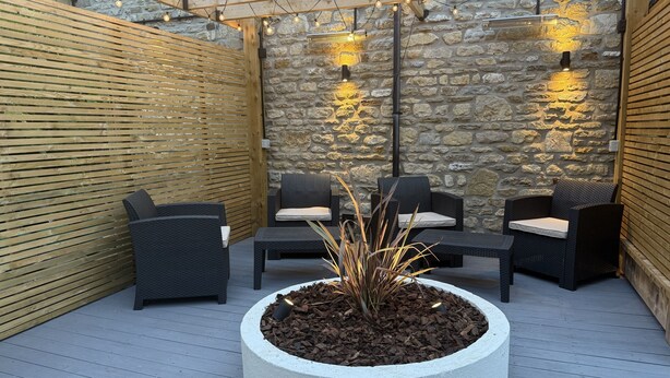 Eine Terrasse im Freien mit Korbmöbeln, einem zentralen Pflanzgefäß mit Pflanzen und Steinmauern mit Beleuchtung.