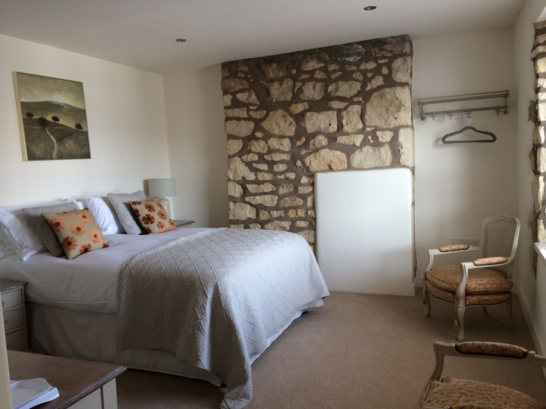 A bedroom with a stone wall, a bed with floral pillows, a bedside table, a chair, and a coat rack.