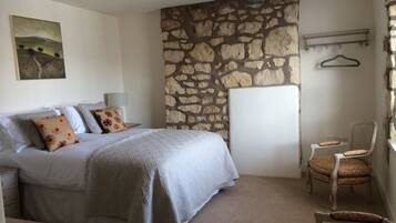 A bedroom with a stone wall, a bed with floral pillows, a bedside table, a chair, and a coat rack.