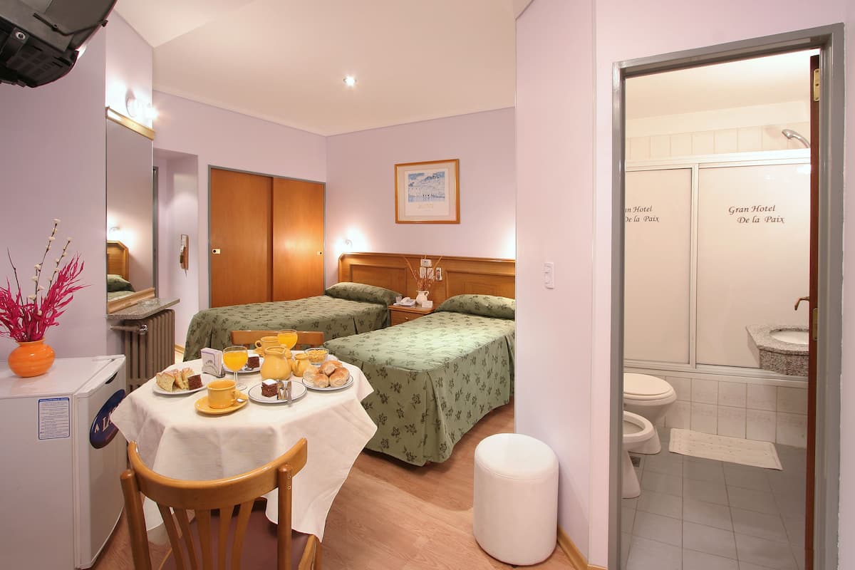 A hotel room with two beds, a dining table with breakfast, a small fridge, and a bathroom with a toilet and sink.