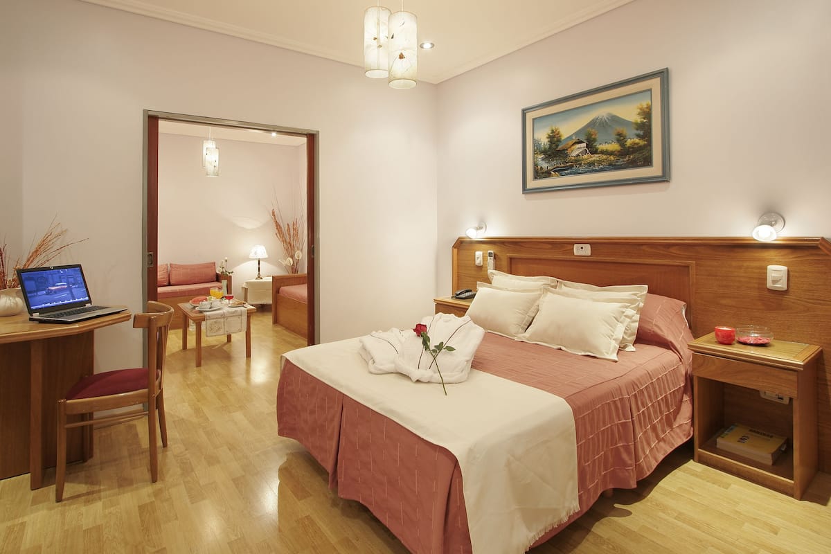 A hotel room with a bed, a desk with a laptop, a chair, a bedside table with a lamp, and a painting on the wall.