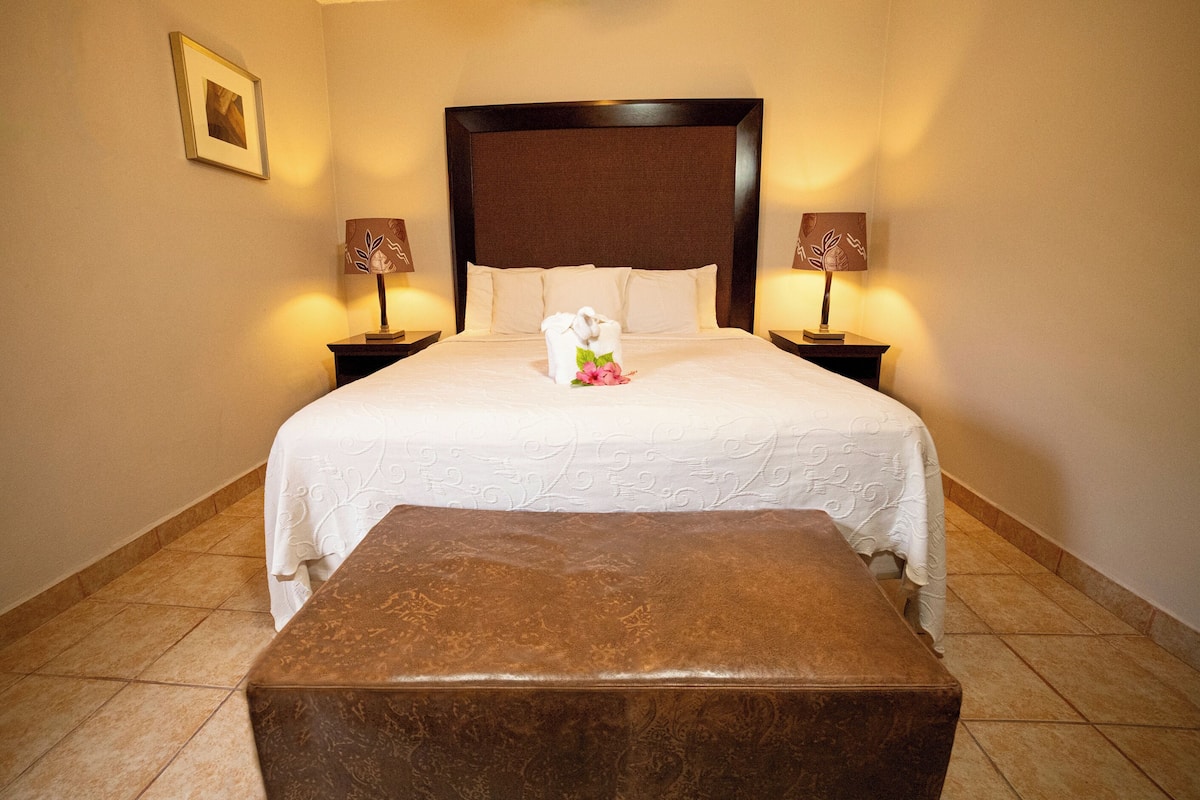 Un lit bien fait, avec un couvre-lit blanc et des oreillers, deux lampes de chevet et un banc marron, dans une chambre d’hôtel.