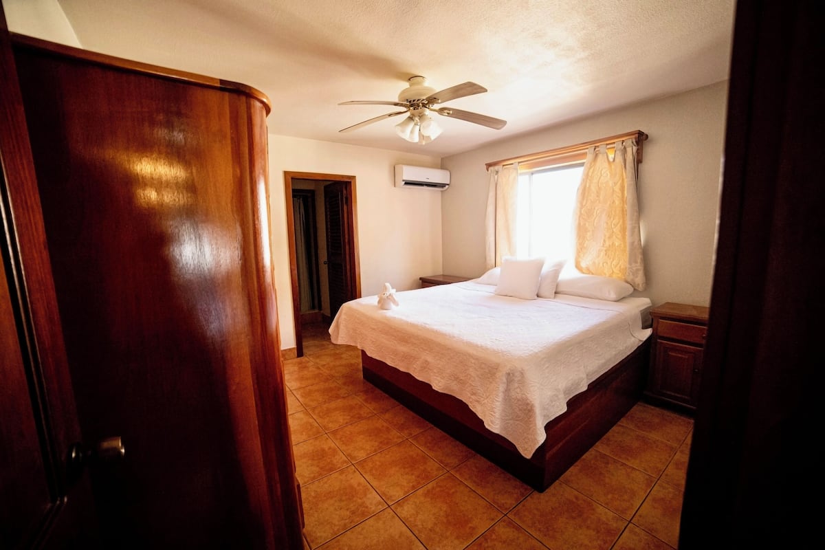 Une chambre à coucher avec un lit en bois, du linge de lit blanc, un ventilateur de plafond et une fenêtre avec des rideaux.
