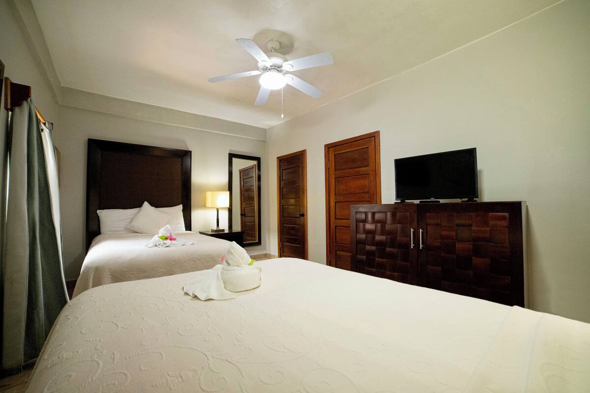 Une chambre d’hôtel avec deux lits, un ventilateur de plafond, un téléviseur posé sur un meuble et des portes en bois.