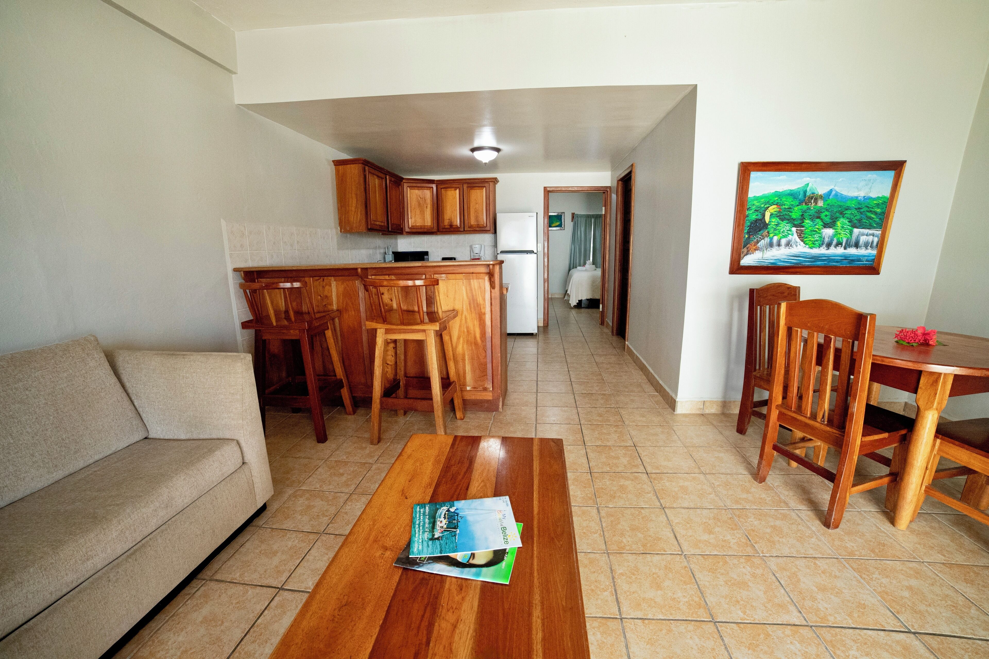 Sala de estar con sofá, mesa de comedor de madera y sillas. Hay una cocina con armarios de madera y un refrigerador. Un cuadro cuelga en la pared, y una puerta da a otra habitación.