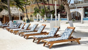 A row of wooden lounge chairs with blue and white patterned cushions on a sandy beach, palm trees, and a building with balconies in the background. - Corona Del Mar (San Pedro)