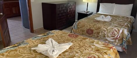 Une chambre d’hôtel avec deux lits, chacun recouvert d’une courtepointe à motifs floraux et muni d’une serviette. On y trouve une armoire en bois et une table de chevet avec une lampe.