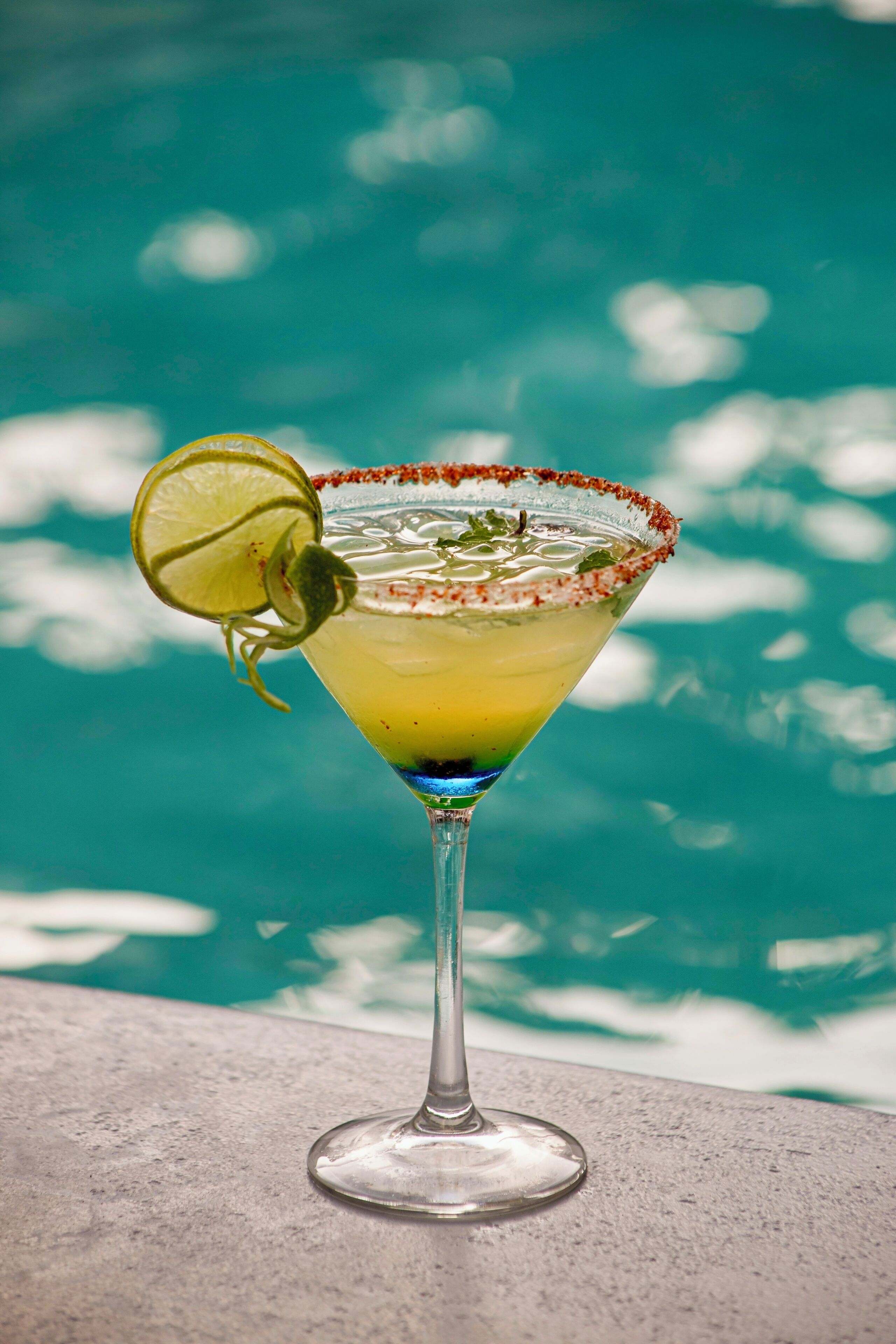 A margarita cocktail with a salted rim, garnished with a lime slice, placed on a concrete surface near a pool.