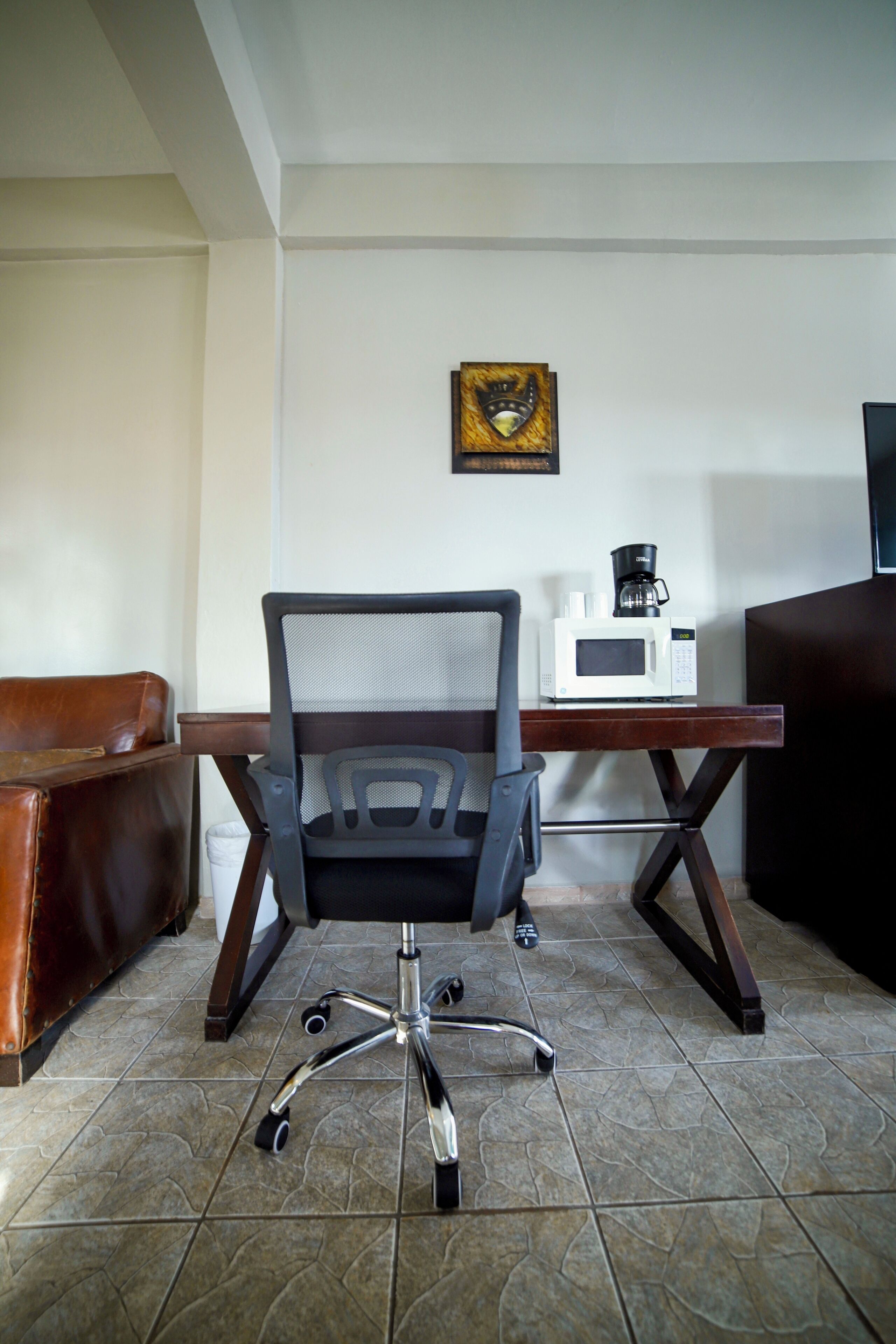 Une pièce avec un fauteuil en cuir marron, un bureau en bois sur lequel se trouve un micro-ondes blanc, et un tableau encadré accroché au mur.