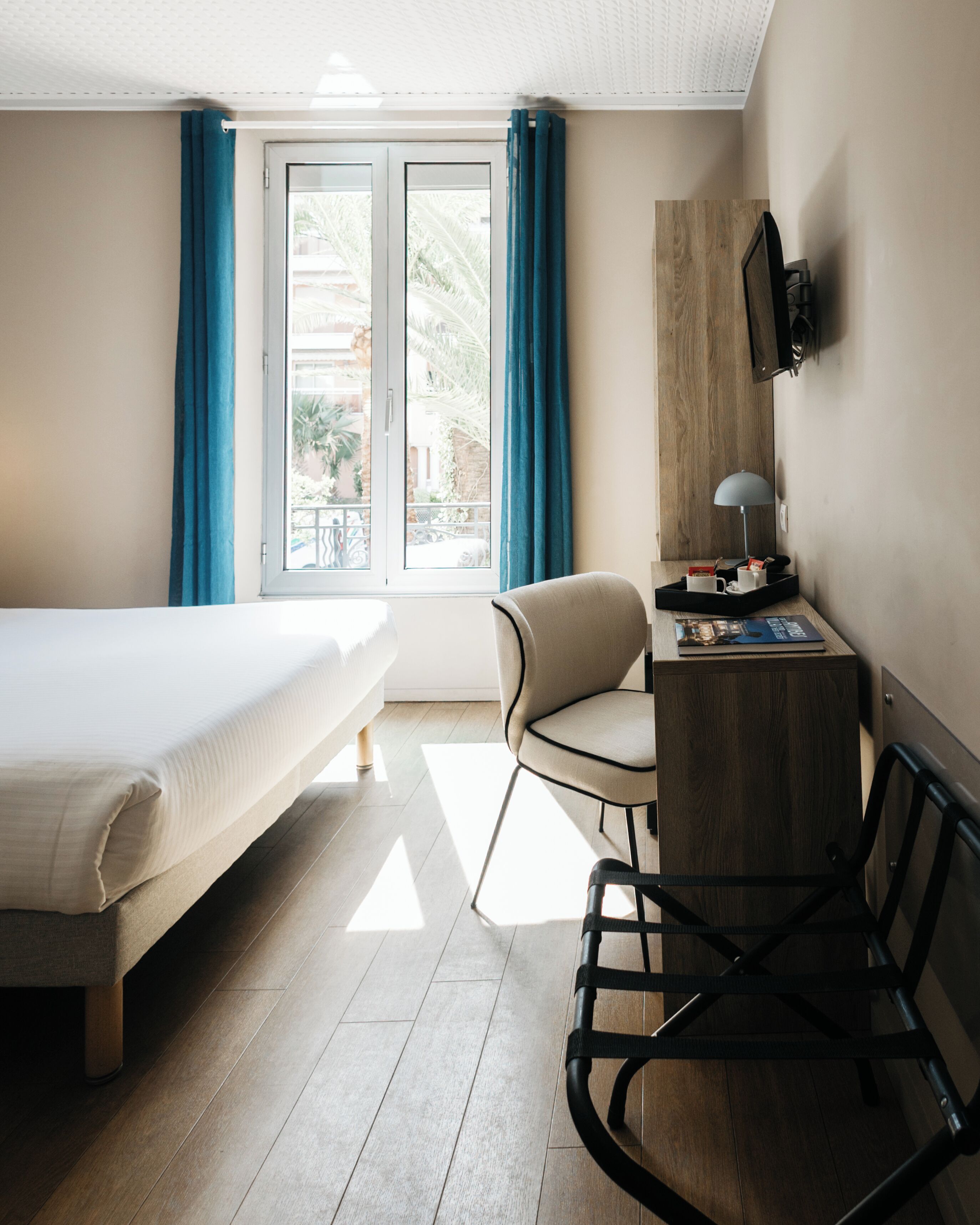 Habitación de hotel con una cama, un escritorio con silla, un televisor montado en la pared y una ventana con cortinas.