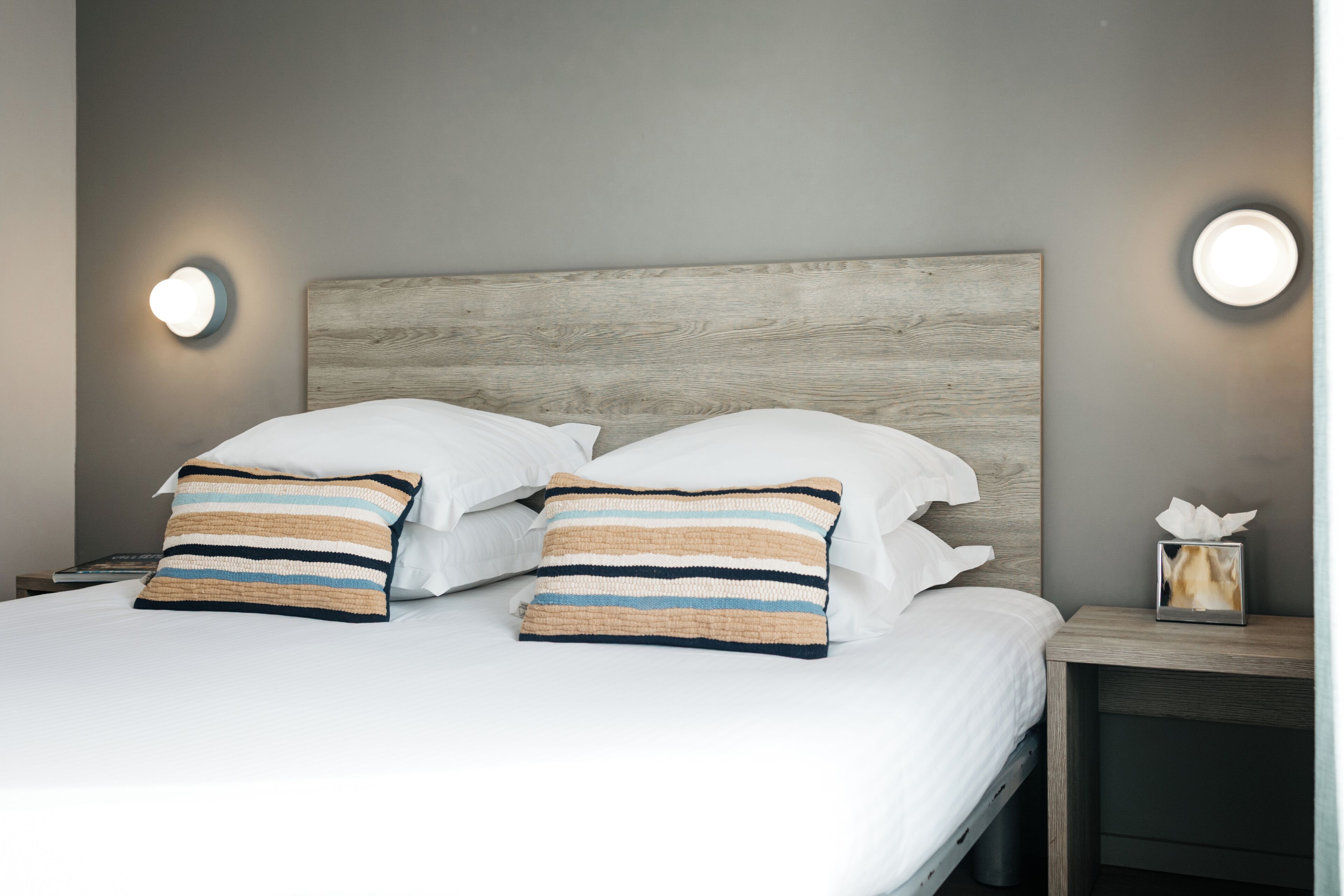 Una cama bien hecha con sábanas blancas, dos almohadas a rayas, un cabecero de madera y lámparas de noche.