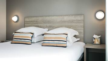 Una cama bien hecha con sábanas blancas, dos almohadas a rayas, un cabecero de madera y lámparas de noche.